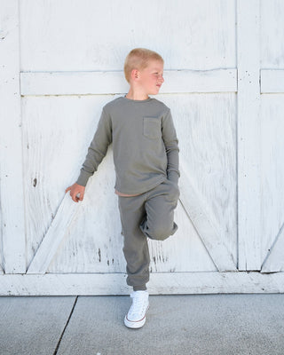 Little Bipsy Waffle Top - Green
A young boy wearing a matching gray long-sleeve shirt and jogger pants stands against a white wooden barn door, with one leg kicked up. He is wearing white sneakers.
