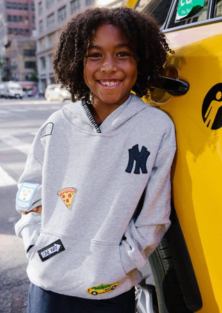 Lola and The Boys All About The Patch City Hoodie
A smiling young girl with curly hair wearing a light gray hoodie with a navy New York Yankees logo and colorful patches, standing next to a yellow taxi in an urban city setting.