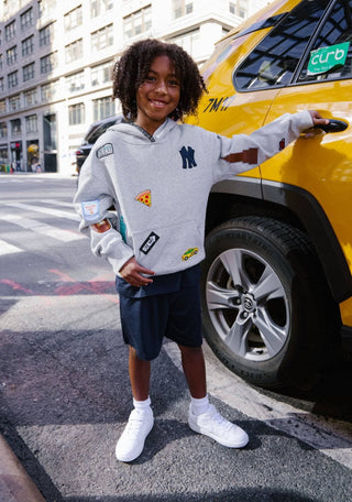 Lola and The Boys All About The Patch City Hoodie
A young boy stands on a city street next to a yellow taxi, wearing a light gray hoodie with multiple colorful patches and a dark navy blue sweatshirt underneath, paired with white sneakers.