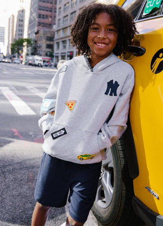 Lola and The Boys All About The Patch City Hoodie
A smiling young boy with curly hair stands on a city street next to a yellow taxi, wearing a light gray hoodie with a navy Yankees logo, a pizza patch, and a small yellow taxi patch, paired with navy shorts.