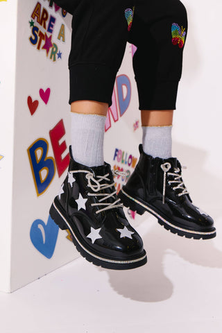 Lola and The Boys Black Rainbow Shimmer Stars Boots
Black patent leather lace-up boots with white star appliqués on the sides, worn with white socks and black pants with colorful patches.