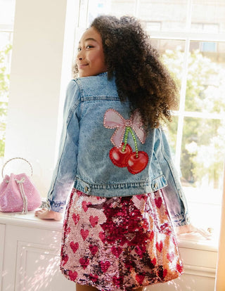 Lola and The Boys Crystal Cherry Bow Denim Jacket
Child wearing a denim jacket with cherry and bow design, standing in a bright room.