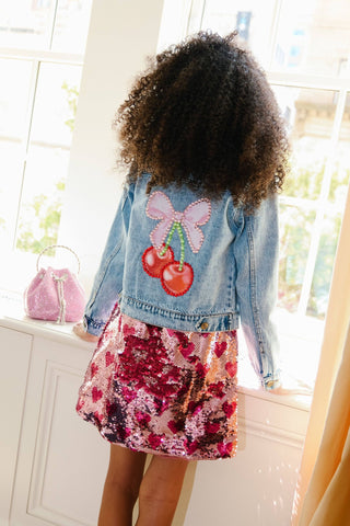Lola and The Boys Crystal Cherry Bow Denim Jacket A child wearing a light blue denim jacket with a pink bow and red cherry patch on the back, paired with a pink and red sequined skirt, standing in front of a bright window.