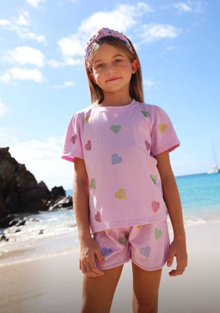 Lola and The Boys Crystal Hearts Short Set - Lavender
Young girl in pink heart-patterned outfit standing on a beach with blue sky and ocean.