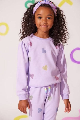 Lola and The Boys Lavender Crystal Hearts Set
A young girl with curly dark hair wearing a light purple two-piece pajama set with glittery heart details, a matching purple headband, and standing against a white background with colorful letter shapes.