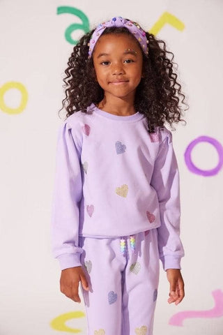 Lola and The Boys Lavender Crystal Hearts Set
Child wearing a purple outfit with heart patterns against a colorful background