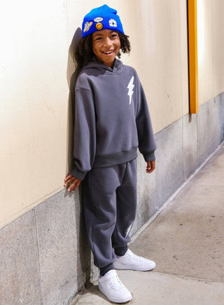 Lola and The Boys Lighting Flash Set - Grey
A smiling child wearing a dark gray hoodie and matching sweatpants with a white lightning bolt graphic, paired with a bright blue beanie and white sneakers, leaning against a beige wall.
