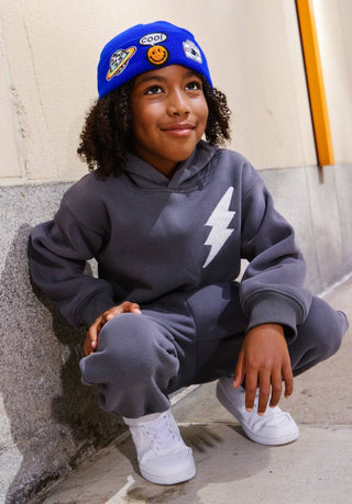 Lola and The Boys Lighting Flash Set - Grey
A child wearing a dark gray matching hoodie and sweatpants set with a white lightning bolt graphic on the chest, crouching against a concrete wall. The child is also wearing a bright blue beanie with cartoon patches and white sneakers.