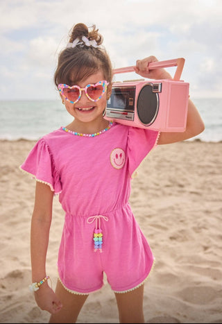 Lola and The Boys Pink Gems Emoji Romper 
Child in pink outfit with heart-shaped sunglasses holding a pink radio on a beach.