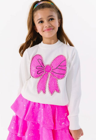 Lola and The Boys Pinky Big Bow Sweater
A young girl wearing a white long-sleeve sweatshirt with a large pink sequined bow on the front, paired with a bright pink sequined ruffled skirt, against a plain white background.