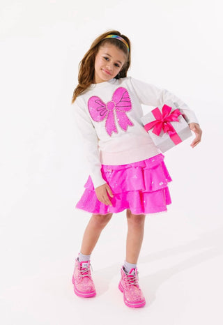 Lola and The Boys Pinky Big Bow Sweater
A young girl modeling a white long-sleeve top with a large pink sequined bow on the front, paired with a bright pink tiered ruffle skirt and pink sneakers, while holding a small gift box with a pink bow against a white studio background.