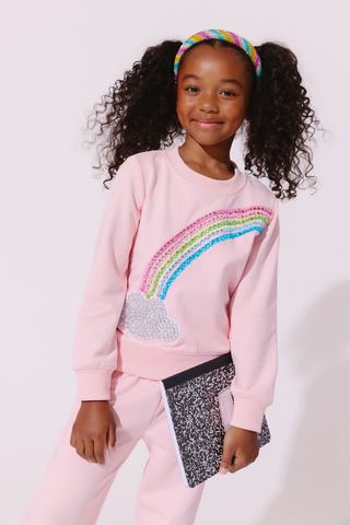 Lola and The Boys Rainbow Dreams Joggers Set
A young girl with curly black hair wearing a pink sweatshirt and matching sweatpants, featuring a colorful sequined rainbow design on the front. She is holding a black and white speckled clutch bag and wearing a rainbow headband against a plain light background.