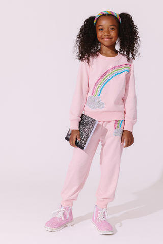 Lola and The Boys Rainbow Dreams Joggers Set
A young girl with curly hair wearing a pink two-piece sweatshirt and jogger set with rainbow and cloud sequin details, holding a glittery notebook, against a light pink studio background.