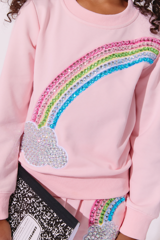 Lola and The Boys Rainbow Dreams Joggers Set
A close-up of a child wearing a light pink sweatshirt with a large rainbow and cloud applique decorated with sparkling rhinestones. The child is also wearing matching pink pants and holding a black-and-white patterned notebook.