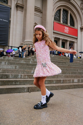 Lola and The Boys Rosie Bows Dress Young girl in a pink dress standing on steps in front of a building with a red sign.