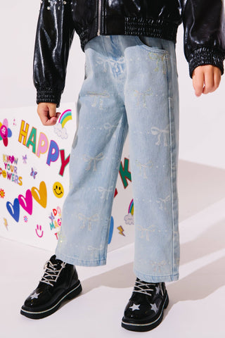 Lola and The Boys Sparkle Bows Denim Jeans
A child wearing light-wash wide-leg denim jeans with embroidered bow details, styled with a black jacket and black star-spangled boots, standing in front of a white wall decorated with colorful stickers.