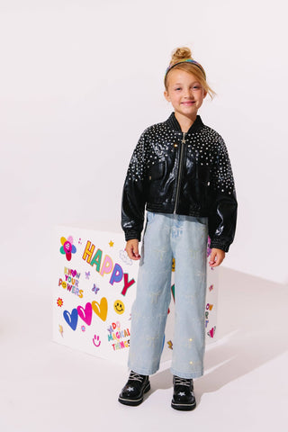 Lola and The Boys Sparkle Bows Denim Jeans
A young girl stands against a white studio background wearing a shiny black bomber jacket covered in silver studs, light blue jeans, and black sneakers. A large white gift bag with colorful 'HAPPY' lettering and doodles stands beside her.