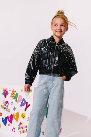 Lola and The Boys Sparkle Bows Denim Jeans
A young girl with blonde hair in a bun stands wearing a shiny black bomber jacket covered in silver studs, paired with light blue jeans. She is posing next to a white box decorated with colorful stickers and the word 'HAPPY'.