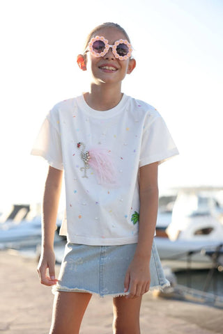 Lola and The Boys Sparkle Flamingo T-shirt
Young girl wearing a white t-shirt with colorful designs and denim shorts, standing outdoors with boats in the background.