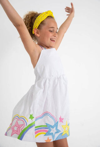 Lola and The Boys Summer Pop Dress
Young girl wearing a white dress with colorful star and rainbow designs, standing against a plain background.
