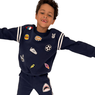 Lola and The Boys Varsity Patch Jogger Set - Navy
A young boy wearing a navy blue sweatshirt and matching sweatpants set, featuring various colorful embroidered patches and white stripe details on the sleeves, posed against a solid white background.