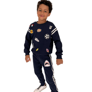 Lola and The Boys Varsity Patch Jogger Set - Navy
A young boy modeling a navy blue tracksuit with various sports-themed patches, including a soccer ball, basketball, and shark graphic, on a solid white background.