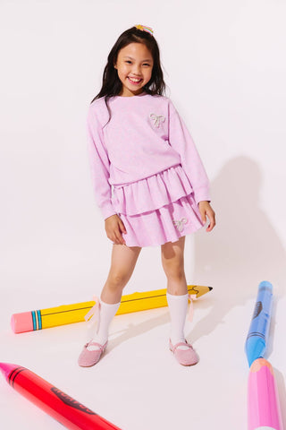 Lola and the Boys Super Soft Pearl Bow Set - Lavender
A young girl stands smiling in a studio wearing a light pink long-sleeve top and matching tiered skirt set. She is styled with white knee-high socks and pink shoes, surrounded by oversized colorful crayons on a white background.