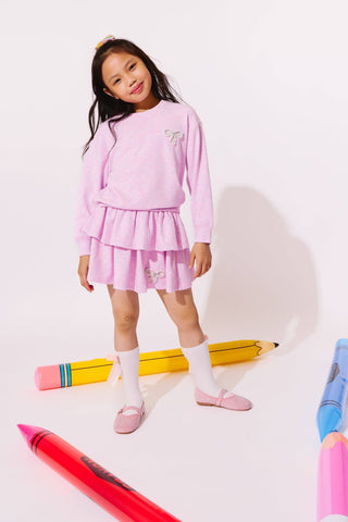 Lola and the Boys Super Soft Pearl Bow Set - Lavender
A young girl stands in a studio wearing a light pink long-sleeve top and matching ruffled skirt, with white socks and pink shoes. Large colorful crayon props are placed around her on a white background.