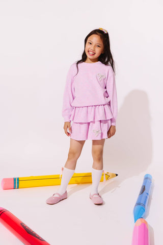 Lola and the Boys Super Soft Pearl Bow Set - Lavender
A young girl stands in a studio wearing a light pink two-piece outfit consisting of a long-sleeve top and a ruffled mini skirt, paired with white knee-high socks and pink shoes. Large colorful toy crayons are arranged on the floor around her against a plain white background.