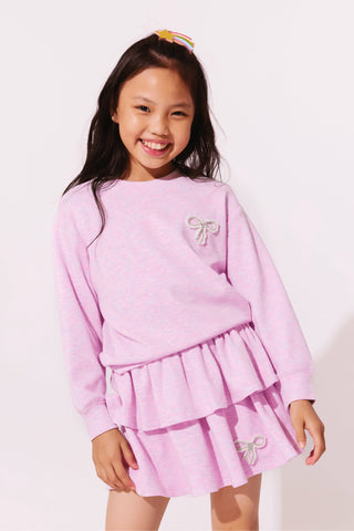 Lola and the Boys Super Soft Pearl Bow Set - Lavender
A young girl smiling and wearing a light pink long-sleeve sweatshirt and matching pleated mini skirt set, both featuring small bow embellishments, against a plain white background.