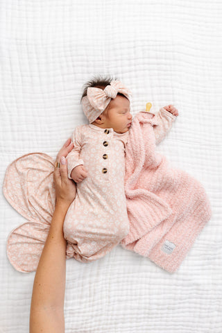 Lou Lou and Company Headband - Louise
A newborn baby wearing a light pink patterned footed romper, matching bow headband, and a soft pink blanket, lying on a white textured blanket with an adult's hand holding the romper.