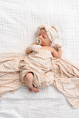 Lou Lou and Company Headband - Milo
A sleeping newborn baby wrapped in a peach and cream gingham muslin swaddle blanket, matching headband, and small matching mittens, lying on a white textured muslin sheet.