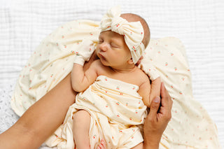 Lou Lou and Company Headband - Sadie
A sleeping newborn baby wrapped in a cream muslin swaddle blanket with tiny red floral prints, wearing a matching cream bow headband and swaddle glove, held by an adult's hands on a white textured blanket.