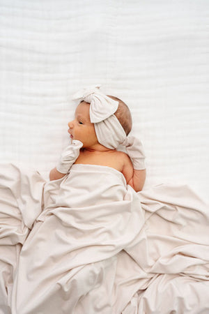 Lou Lou and Company Headband - Winnie
A sleeping newborn baby wrapped in a soft cream-colored muslin swaddle blanket, wearing a matching cream bow headband and cream mittens, lying on a textured white blanket.