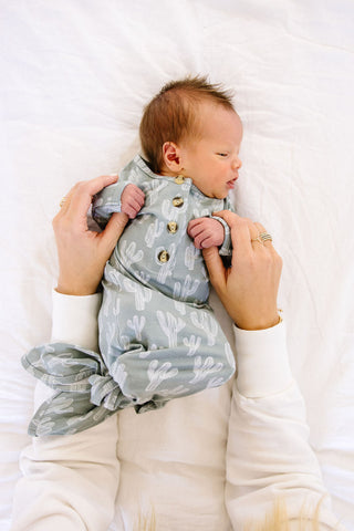 Lou Lou and Company Knotted Gown - Knox
A newborn baby wearing a sage green and white cactus print romper, held by an adult's hands against a white fabric background.
