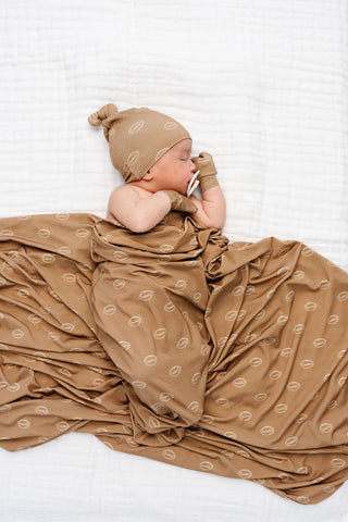 Lou Lou and Company Swaddle Blanket - Brady
A newborn baby lying on a white textured blanket, wrapped in a brown muslin swaddle blanket with a repeating circular pattern, wearing a matching brown knit bonnet and glove.