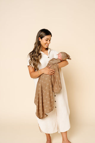 Lou Lou and Company Swaddle Blanket - Brady
A woman in a white button-front dress holds a baby wrapped in a taupe patterned blanket against a solid beige studio background.