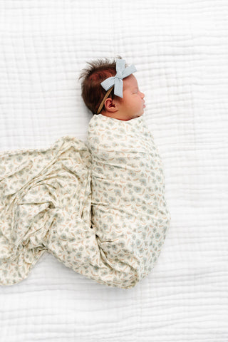 Lou Lou and Company Swaddle Blanket - Faye
A newborn baby lying on a white textured blanket, swaddled in a cream-colored muslin swaddle blanket with a delicate green and beige floral pattern, and wearing a matching light blue bow headband.