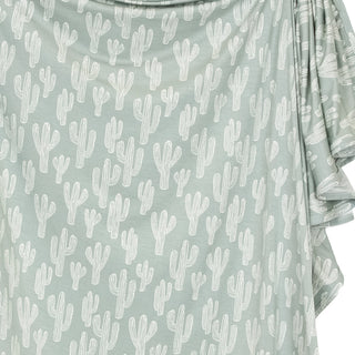 Lou Lou and Company Swaddle Blanket - Knox
A close-up of a draped fabric top in sage green with an all-over white cactus print, featuring ruffled side details.