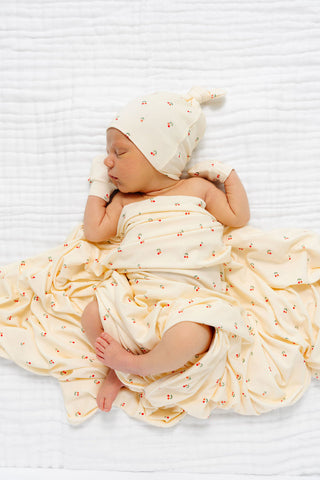 Lou Lou and Company Swaddle Blanket - Sadie
A sleeping newborn baby wrapped in a cream-colored muslin swaddle blanket with a tiny red and orange floral pattern, wearing a matching beanie hat, lying on a white textured blanket.