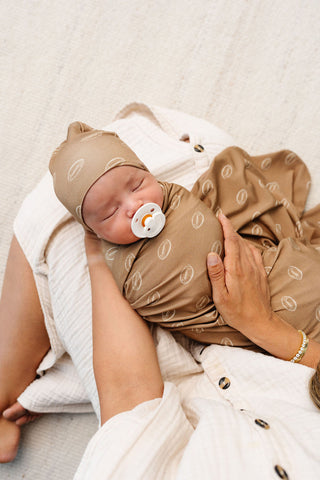 Lou Lou and Company Top Knot Hat - Brady
A sleeping newborn baby swaddled in a brown muslin swaddle blanket with a matching bonnet, resting on a mother's lap. The swaddle and bonnet feature a light beige circular pattern, and the baby is wearing a white muslin dress with gold buttons.