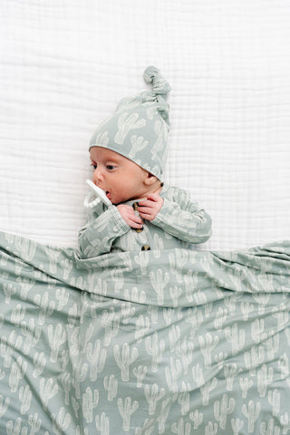 Lou Lou and Company Top Knot Hat - Knox
Baby wrapped in a green and white patterned swaddle blanket with a matching hat.