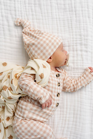 Lou Lou and Company Top Knot Hat - Milo
A sleeping newborn baby wearing a peach and cream gingham long-sleeve romper, matching pointed hat, and a cream blanket with teddy bear prints, lying on a white quilted blanket.