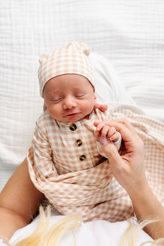 Lou Lou and Company Top Knot Hat - Milo
A sleeping newborn baby wrapped in a soft beige and white gingham swaddle blanket and matching hat, held by an adult's hand against a white textured background.