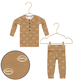 Lou Lou and Company Top and Bottoms - Brady
A toddler's two-piece pajama set in a warm tan color, featuring a long-sleeve henley top and matching pants, both printed with a repeating football pattern. The set is displayed on gold-colored hangers against a white background, with a circular close-up inset showing the fabric texture and football print detail.