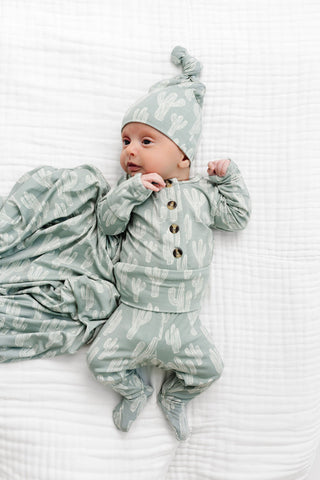 Lou Lou and Company Top and Bottoms - Knox
A baby lying on a white muslin blanket wearing a sage green and white cactus print footed sleeper with a matching beanie and a matching cactus print swaddle blanket.