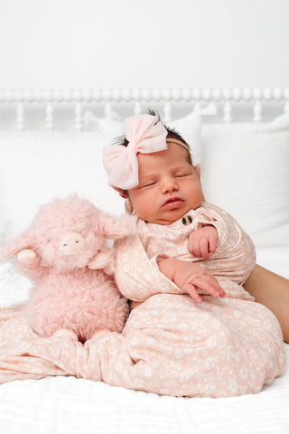 Lou Lou and Company Top and Bottoms - Louise
A sleeping newborn baby wrapped in a light pink patterned swaddle blanket, wearing a matching pink bow headband, and cuddling a fluffy pink stuffed sheep on a white bed.