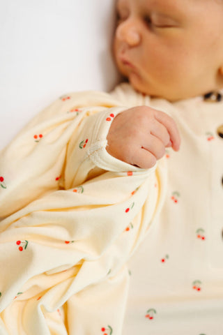 Lou Lou and Company Top and Bottoms - Sadie
A close-up of a sleeping newborn baby wearing a cream-colored long-sleeve bodysuit and swaddle blanket with a small red cherry and green leaf pattern.