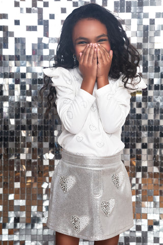MIA New York Heart Skort - Silver
A young girl with long, curly black hair stands in front of a silver mirror mosaic wall, wearing a white long-sleeve top with embroidered hearts and a silver skirt with heart-shaped rhinestone embellishments.