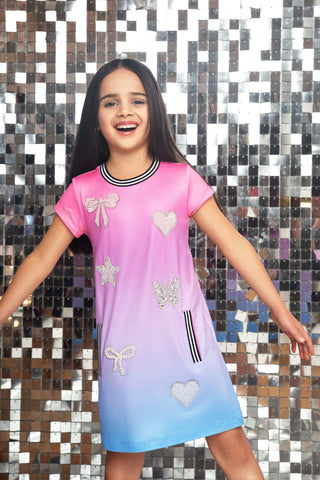 MIA New York Icon Dress - Purple Ombre
A young girl smiling and posing in a short-sleeve gradient dress that transitions from pink at the top to light purple and blue at the bottom, decorated with glittery bows, hearts, and stars, set against a silver mirror mosaic wall.